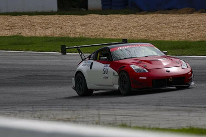 A red and white race car with the number 50 on the door speeds around a racetrack, equipped with a 9livesracing 350z Big Wang Kit '02-08 Z33, featuring GT3 performance and extruded aluminum wings. The car boasts a prominent rear spoiler and various sponsor logos. The track is lined with grass and barriers, with tire walls and gravel in the background.