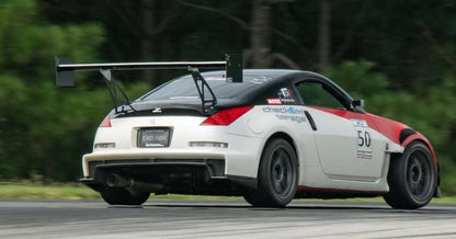 The 350z Big Wang Kit '02-08 Z33, by 9livesracing, transformed a modified sports car bearing the number 50 on its door and equipped with a substantial rear wing crafted from extruded aluminum wings, decals, and a wide body kit. The sleek white car is accented in red and black as it charges around the racetrack. Blurred greenery in the background signals high speed, highlighting its impressive Big Wang GT3 performance.