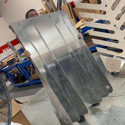 A person is partially hidden behind a large, metallic Miata Diffuser '90-05 NA/NB from 9livesracing, holding it up with both hands. The workshop-like environment contains various tools, materials, and a partially visible wooden structure in the background. The aluminum Miata Diffuser '90-05 NA/NB indicates an ongoing car modification project.