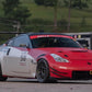 A modified red and silver Nissan 350Z equipped with a 9livesracing 350z Big Wang Kit '02-08 Z33 performance wing and an aftermarket front bumper is parked on a tarmac. The car is adorned with several racing stickers, including one featuring the number 50 on the door. Trees and a trailer are visible in the background.