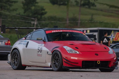 A modified red and silver Nissan 350Z equipped with a 9livesracing 350z Big Wang Kit '02-08 Z33 performance wing and an aftermarket front bumper is parked on a tarmac. The car is adorned with several racing stickers, including one featuring the number 50 on the door. Trees and a trailer are visible in the background.