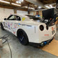 A white sports car adorned with various racing stickers and decals sits in a garage. The car features a GT-R Big Wang Kit '07-22 from 9livesracing, along with down-force adjustments, blue alloy wheels, and a prominent exhaust system. The garage background shows tools and equipment scattered around.