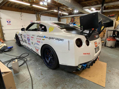 A white sports car adorned with various racing stickers and decals sits in a garage. The car features a GT-R Big Wang Kit '07-22 from 9livesracing, along with down-force adjustments, blue alloy wheels, and a prominent exhaust system. The garage background shows tools and equipment scattered around.