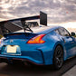 A vivid blue 370z Big Wang Kit '08-20 Z34 from 9livesracing, featuring extruded aluminum wings and a large rear spoiler, is parked on a suburban street at sunset. The car's taillights are illuminated, while dark clouds in the sky are partially lit by the setting sun. Houses and street lights can be seen in the background.