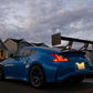 A sleek blue sports car, featuring the 370z Big Wang Kit '08-20 Z34 by 9livesracing with extruded aluminum wings for optimal down-force adjustments, is parked on a suburban street in front of houses. The sky shows a mix of clouds with a hint of sunset, creating a dramatic backdrop. The car's license plate is illuminated in the evening light.