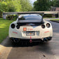 A white sports car with a large rear spoiler, specifically featuring the 9livesracing GT-R Big Wang Kit '07-22 for optimal down-force adjustments, parked on a driveway. The car has several decals on the back, including "Yokohama," "Tire Rack," and "G-LOC." The license plate reads "RC72896." Houses and trees dot the background.