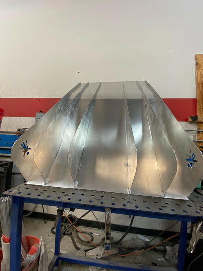 A partially constructed aluminum ventilation unit with several connected panels rests on a perforated workbench. The background shows a red and white wall with various tools and equipment scattered. Electrical cords and other workshop items, including bits from a 9livesracing Miata Diffuser '90-05 NA/NB project, are visible on the floor.
