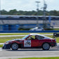 A red and black V8 Miata with number 96 on the door speeds along a race track, featuring a Miata Diffuser '90-05 NA/NB by 9livesracing for enhanced aerodynamics. The background is blurred, indicating motion, and shows racetrack facilities and surrounding landscape under a clear sky. The driver is seen wearing a helmet and racing gear.