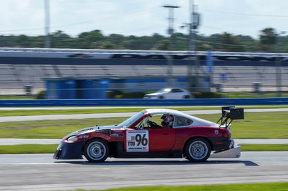 A red and black V8 Miata with number 96 on the door speeds along a race track, featuring a Miata Diffuser '90-05 NA/NB by 9livesracing for enhanced aerodynamics. The background is blurred, indicating motion, and shows racetrack facilities and surrounding landscape under a clear sky. The driver is seen wearing a helmet and racing gear.