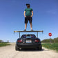 A man wearing a green shirt, black shorts, and a hat stands confidently with his hands in his pockets on the large rear wing of a black sports car featuring a Miata Big Wang Kit '90-05 NA/NB from 9livesracing on a dirt road. A "STOP" sign is visible to the right, and the sky is clear and blue.