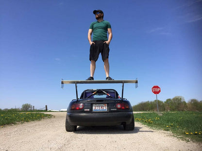 A man wearing a green shirt, black shorts, and a hat stands confidently with his hands in his pockets on the large rear wing of a black sports car featuring a Miata Big Wang Kit '90-05 NA/NB from 9livesracing on a dirt road. A "STOP" sign is visible to the right, and the sky is clear and blue.