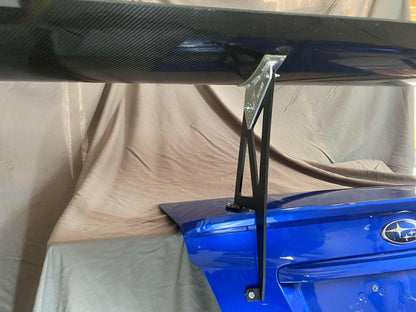 Close-up of the trunk of a blue Subaru Impreza equipped with the Subaru Impreza / WRX / STI Big Wang kit 14-21 VA by 9livesracing. The large carbon fiber spoiler, mounted on sturdy metal brackets for optimal downforce, stands out. Gray drapes form the background, highlighting the prominently visible Subaru logo on the trunk.