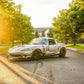A silver race car with the number 615, outfitted with a 9livesracing Miata Big Wang Kit '90-05 NA/NB for added down-force, rear spoiler, front splitter, and various decals, is parked on a street lined with trees and houses in the background. The sun is setting, casting a warm glow over the scene.
