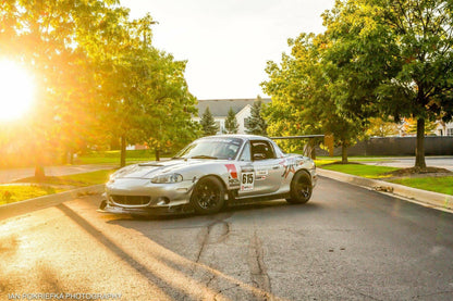 A silver race car with the number 615, outfitted with a 9livesracing Miata Big Wang Kit '90-05 NA/NB for added down-force, rear spoiler, front splitter, and various decals, is parked on a street lined with trees and houses in the background. The sun is setting, casting a warm glow over the scene.