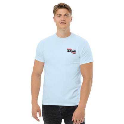 A person with short hair is wearing a light blue Men's Soda Can Wang t-shirt from Nine Lives Racing, made from 100% cotton. The tee features the text "NINE LIVES" and a small graphic on the upper left chest area. They are also wearing black pants, standing against a plain white background, and smiling slightly.