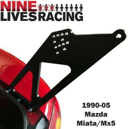 A Miata Big Wang Kit '90-05 NA/NB bracket from 9livesracing, American-made for 1990-2005 Mazda Miata/MX-5 vehicles, mounted on a red surface. This black metal bracket features multiple drilled holes and geometric cutouts, perfect for enhancing downforce with your Mazda Miata.