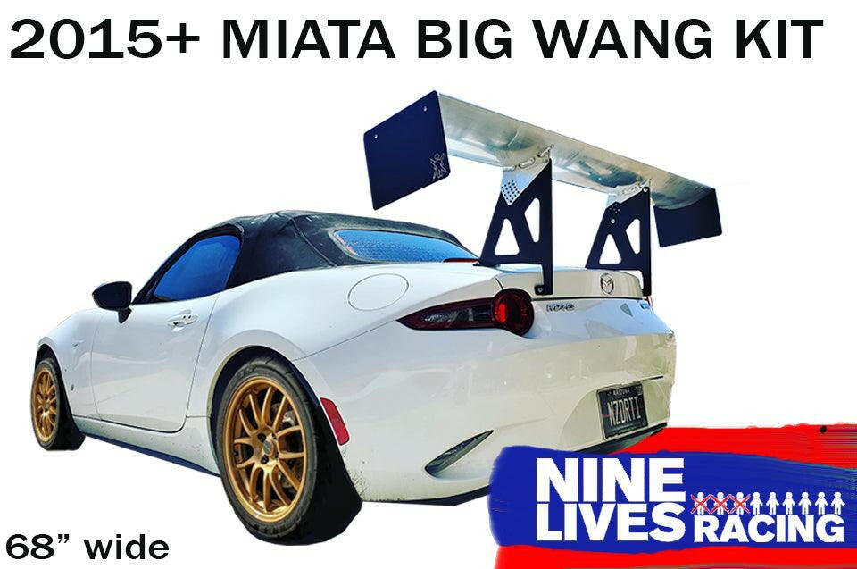 A white convertible car with a black soft top is fitted with an aftermarket high down-force wing on the rear. The text reads "Miata Big Wang Kit 2016+ ND" and "9livesracing." The car, featuring gold-colored wheels and a brand logo in the bottom right corner, boasts an aluminum airfoil for enhanced performance.
