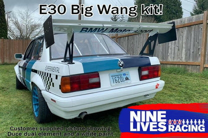 A white 325 BMW race car (’82-94 E30) featuring the 9livesracing Big Wang kit is parked on grass. It has a large rear wing known for its downforce capabilities and sports a "BMW Motorsport" decal on the rear window along with a racing livery. The text "325 Big Wang kit!" is at the top, and the Nine Lives Racing logo with accompanying text is positioned at the bottom right corner.