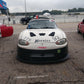 A racing car featuring a sleek white and black design and the number 92 alongside "Hoosier" on the hood is parked on a race track, fitted with the Miata Medium Downforce Kit '90-05 NA/NB from 9livesracing. The driver’s side door displays the number 96, and there is a helmet resting atop the car while other race cars are parked nearby.