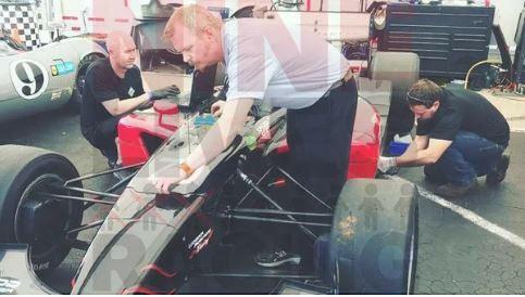 Three men work on a race car in a garage. An aerodynamics expert stands checking the front of the car, utilizing the 9livesracing CFD Wind Tunnel equipment, another kneels working on the rear, and the third sits by another car in the background. The garage has various tools and equipment scattered around.
