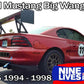 A red Ford Mustang from the 1994-1998 SN95 series is parked on asphalt, featuring a large extruded aluminum wing from the 9livesracing Mustang Big Wang Kit '94-'98 SN95. The car sports race decals including the number 11 and aftermarket wheels. Text reads "Mustang Big Wang GT3" and "9livesracing," highlighting its impressive lift to drag ratio.