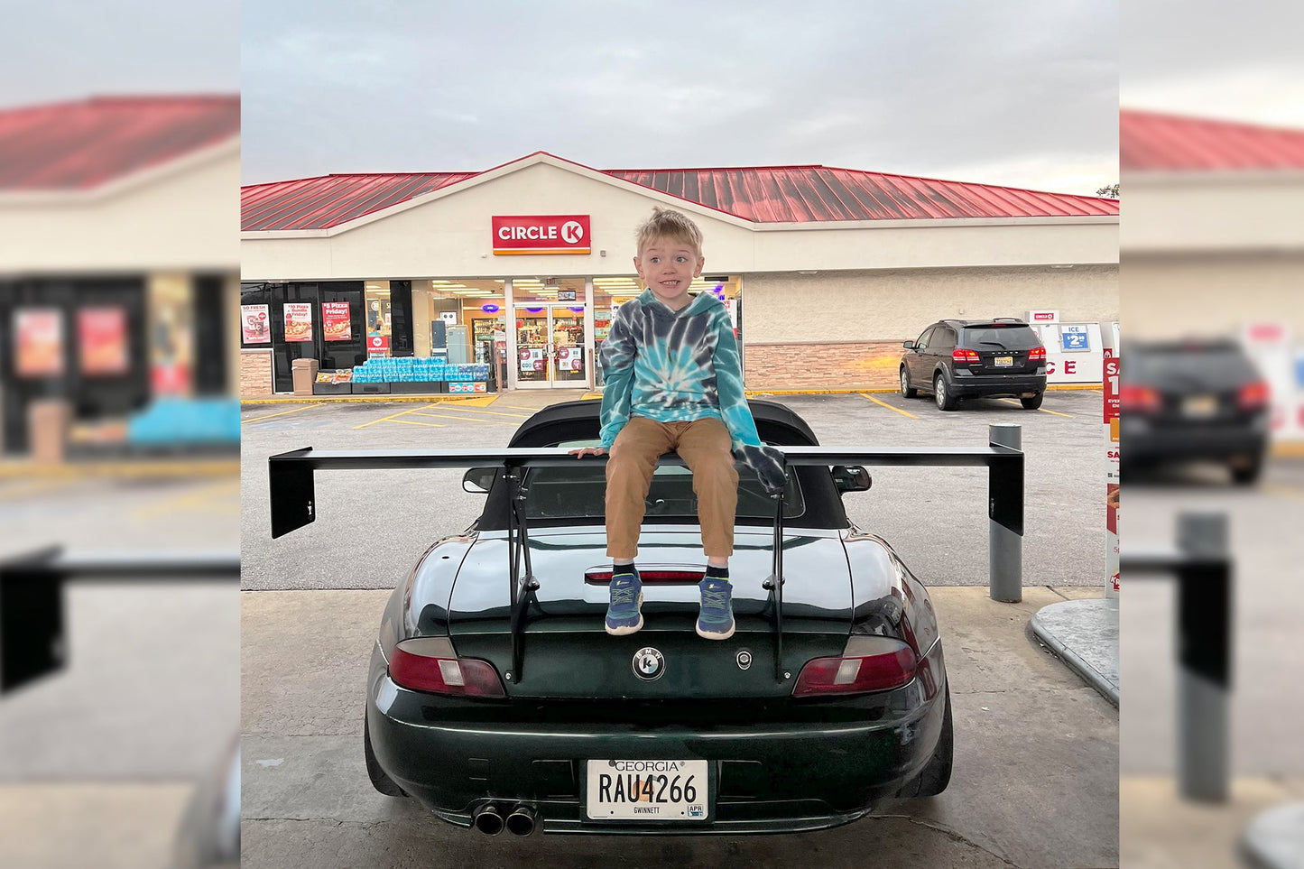 A young boy in a tie-dye hoodie sits on the rear wing of a black sports car at a gas station. Behind him, the Circle K convenience store with a red roof is visible. The gas station forecourt has a few other cars parked in front. Featuring 9livesracing BMW z3 roadster wings, the license plate reads "RAU4266.