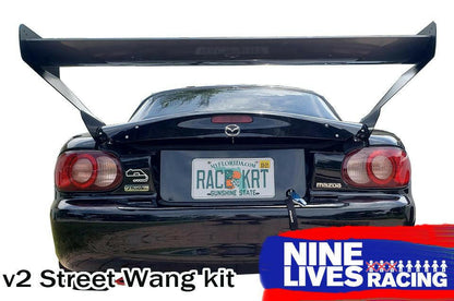 A rear view of a black Mazda features a prominently attached Miata V2 Street Wang '90-05 NA/NB measuring 48 inches. The Florida license plate reads "RAC3KRT," with visible stickers and a rear tow hook. The text at the bottom reads "v2 Street Wang kit" alongside the logo for 9livesracing, emphasizing its performance edge.