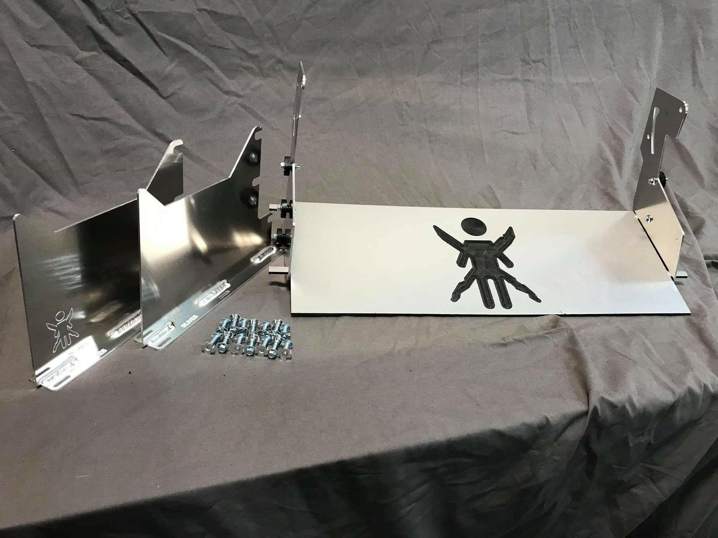 A metal object with multiple parts, screws, and bolts is arranged on a fabric background. The main piece features an engraved design resembling a stylized figure with arms and legs outstretched. Other components include brackets, side panels, and the Miata Sturdy Boii Splitter Mounts '90-05 NA/NB by 9livesracing for increased downforce.