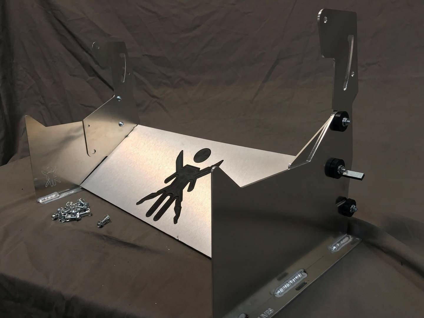 The 9livesracing Miata Sturdy Boii Splitter Mounts '90-05 NA/NB feature a custom metal bracket with a stylized figure cutout design on its front panel, set against a dark backdrop. Resembling Miata Splitter mounts, the bracket includes angled supports on both sides and comes with fasteners placed beside it, ready for assembly.