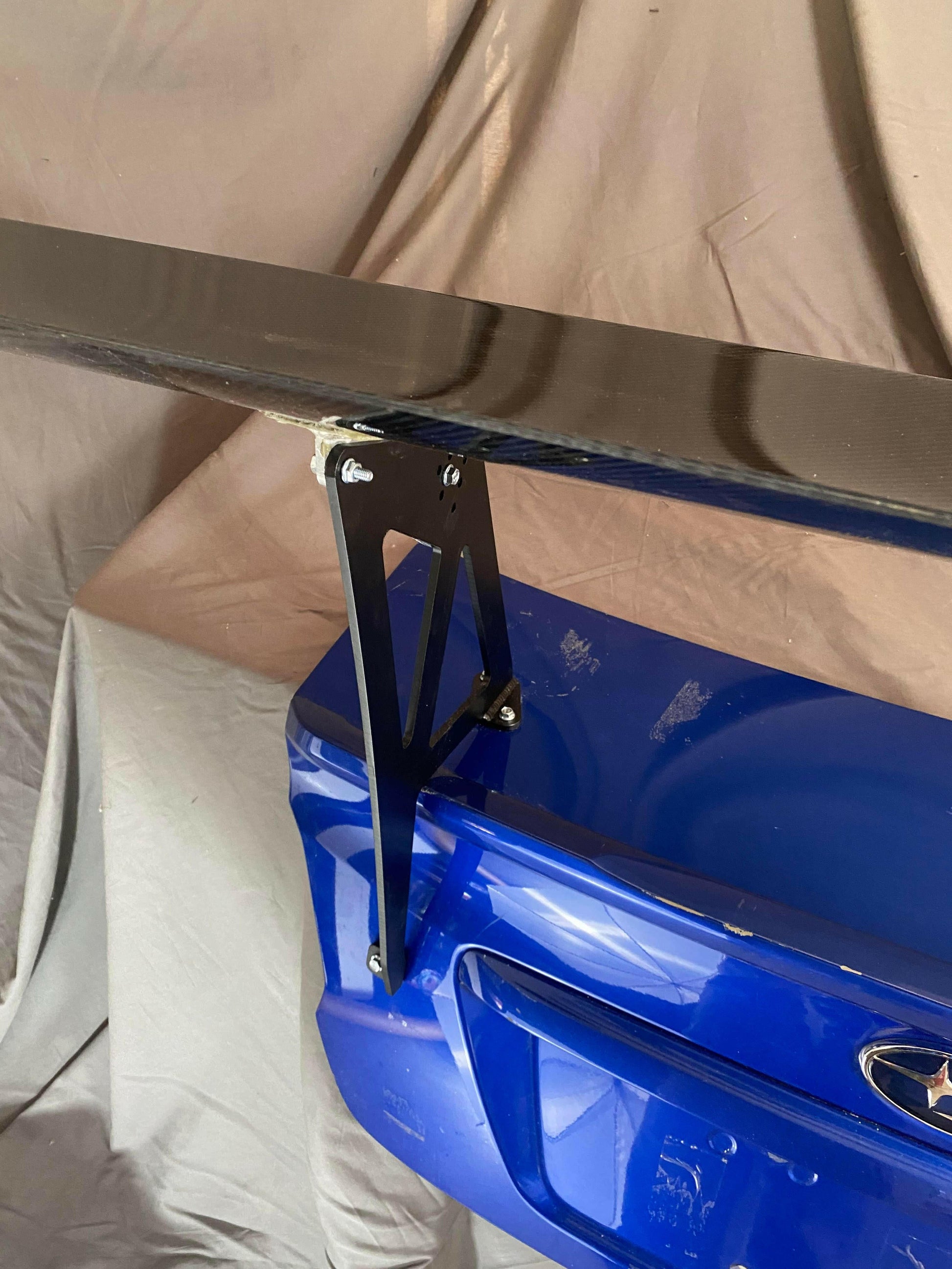 A close-up image of a Subaru Impreza featuring the 9livesracing Big Wang kit (14-21 VA) shows the glossy black rear spoiler designed for optimal downforce, securely mounted on a metal bracket attached to the trunk. The background showcases light-colored fabric draped behind the car.