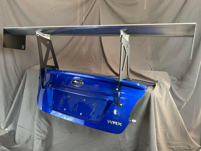 A Subaru Impreza / WRX / STI Big Wang Kit 14-21 VA from 9livesracing, featuring a blue car trunk lid with a large, silver aftermarket aluminum wing attached. The trunk lid proudly displays the Subaru emblem and "WRX" badge. Designed for optimal downforce, this setup is showcased against a gray fabric backdrop.