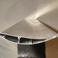 Close-up view of a metal object with a teardrop-shaped cross-section, mounted on a black stand. The surface appears smooth and reflective, with internal compartments and channels visible. This 9livesracing GT-R Big Wang Kit '07-22 features extruded aluminum wings for precise down-force adjustments against the softly lit neutral background.