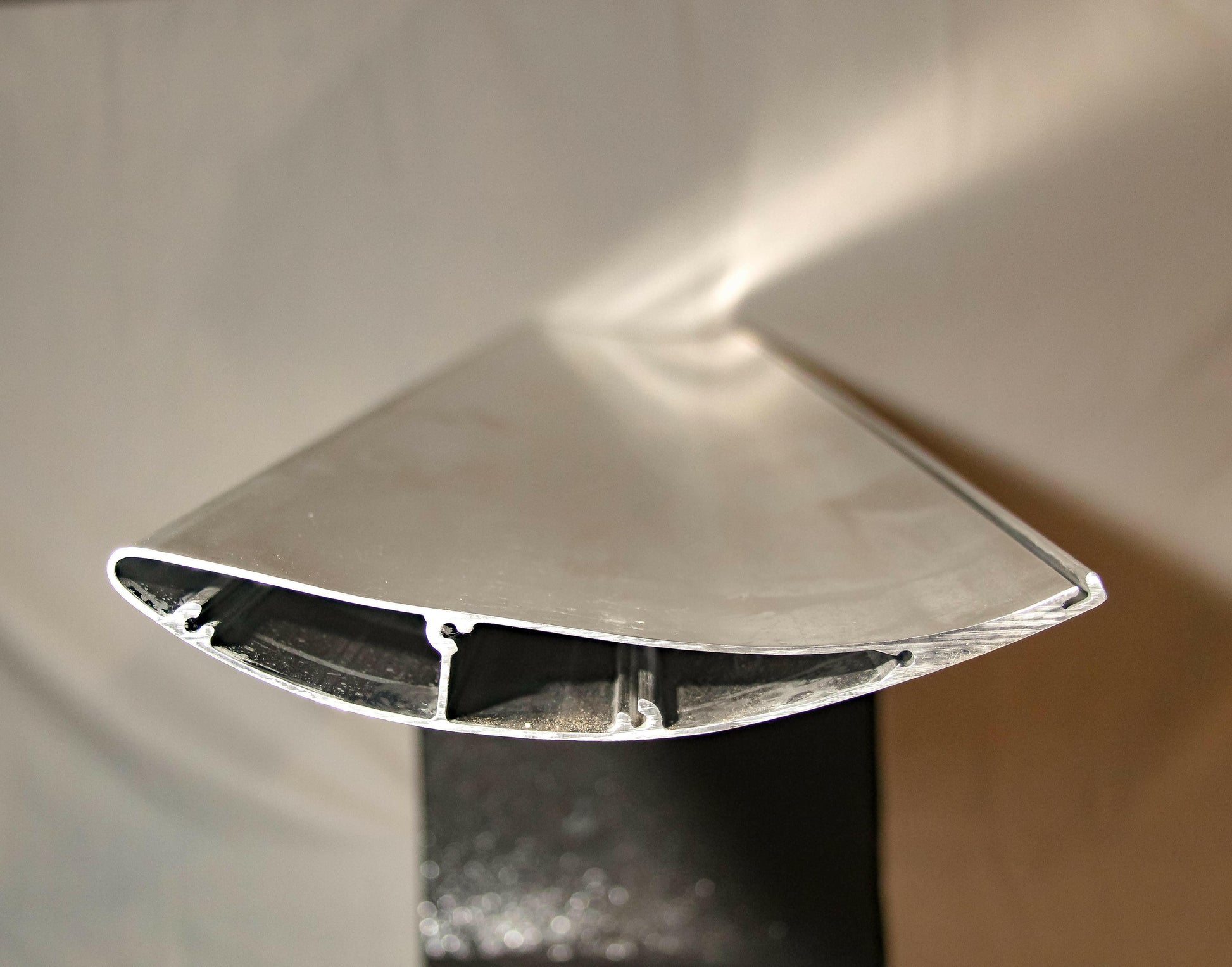 Close-up view of a metal object with a teardrop-shaped cross-section, mounted on a black stand. The surface appears smooth and reflective, with internal compartments and channels visible. This 9livesracing GT-R Big Wang Kit '07-22 features extruded aluminum wings for precise down-force adjustments against the softly lit neutral background.