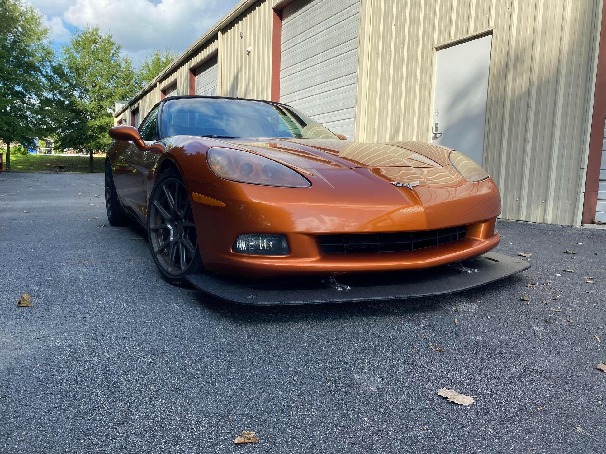 A metallic orange sports car with a sleek design, featuring the Corvette Splitter - C6 & C5 ‘05- 13 by 9livesracing, is parked outside a beige metal building with a white door and garage doors in the background. The car, showcasing CNC cut pockets and carbon elements, is situated on a paved surface with a few fallen leaves scattered on the ground.