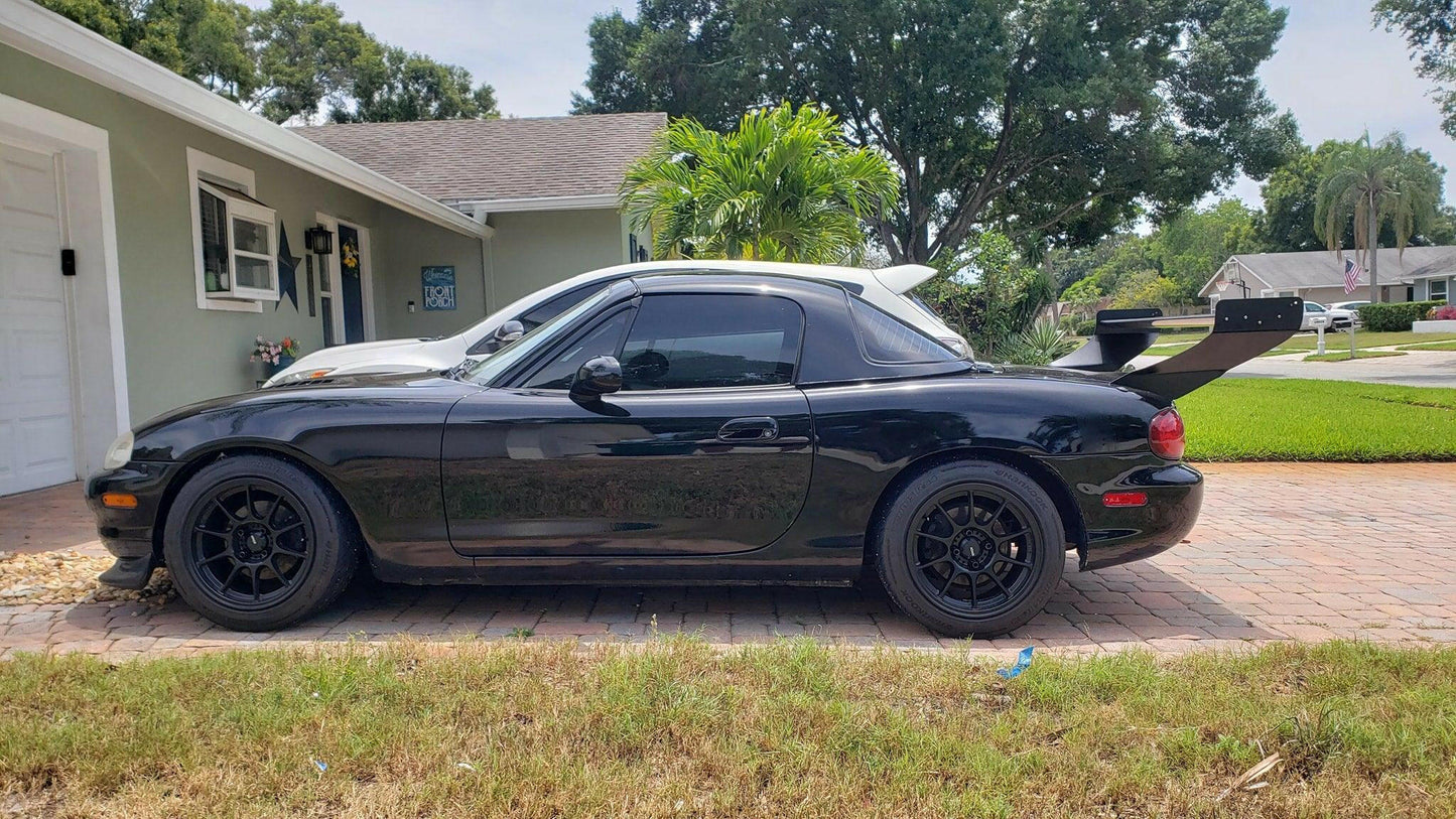 A sleek black convertible sports car, featuring black rims and a Miata V2 Street Wang '90-05 NA/NB from 9livesracing, is parked on a brick driveway in front of a light green house with white trim. Lush greenery and palm trees can be seen in the background on this sunny day, highlighting its performance potential.
