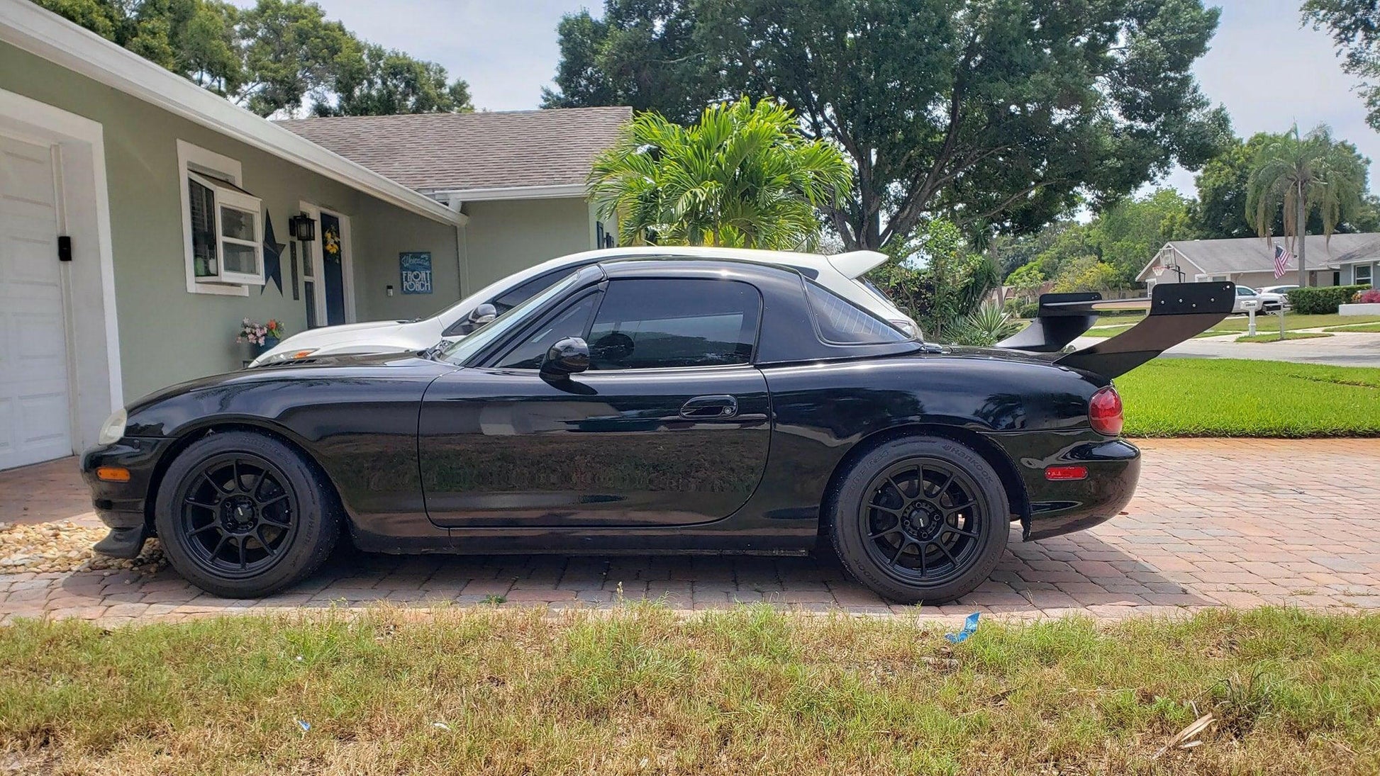 A sleek black convertible sports car, featuring black rims and a Miata V2 Street Wang '90-05 NA/NB from 9livesracing, is parked on a brick driveway in front of a light green house with white trim. Lush greenery and palm trees can be seen in the background on this sunny day, highlighting its performance potential.