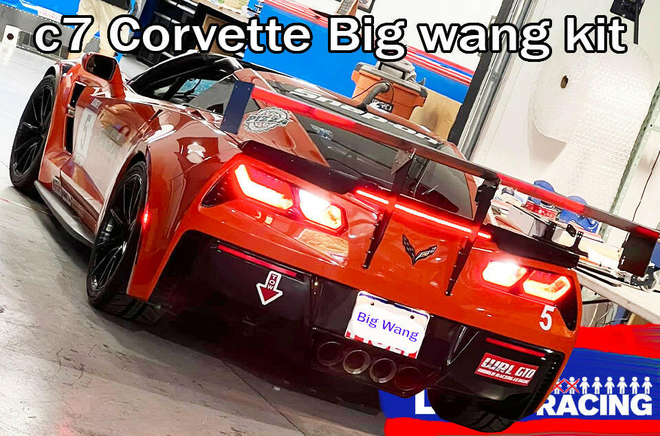 An eye-catching orange Chevrolet Corvette C7 sports car with a prominent rear wing is parked indoors. The license plate reads "Big Wang," amidst visible racing paraphernalia. A sign above the car reads "Corvette Big Wang Chassis Mount ’2014 - 2019 C7 by 9livesracing," emphasizing the aluminum wings designed for optimal down-force adjustments.