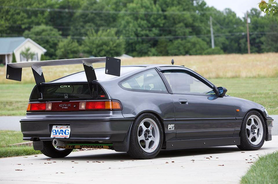A customized gray Honda CRX equipped with the 9livesracing '83-91 CRX Big Wang Kit and aftermarket wheels is parked on a driveway. The car features a lowered stance and several performance modifications, including a rear diffuser and side skirts for enhanced down-force. Trees and grass can be seen in the background.