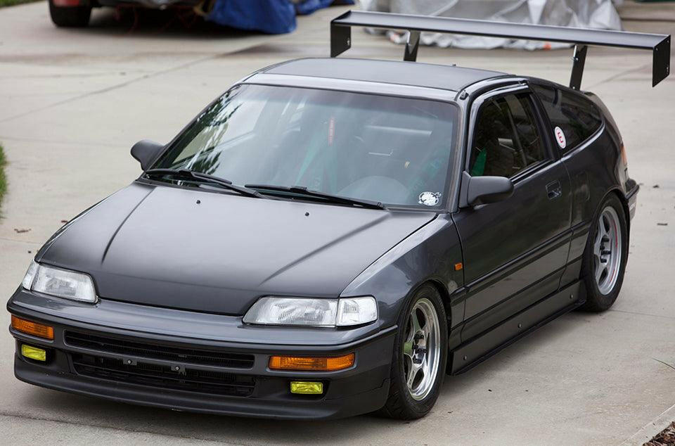 A modified Honda CRX, fitted with the 9livesracing CRX Big Wang Kit '83-91 in matte grey, featuring aerodynamic enhancements such as large aluminum wings and a front splitter to maximize down-force. The car sits low on aftermarket wheels and boasts a clean, polished appearance, parked on a concrete driveway.