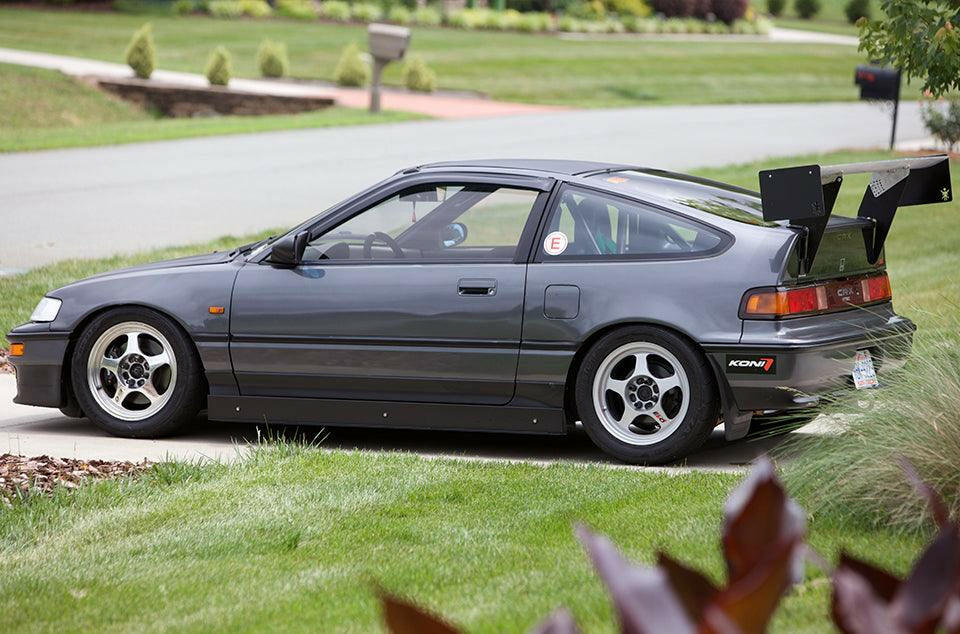 A gray Honda CRX with a sporty appearance is parked on a driveway, featuring modified elements such as 9livesracing's CRX Big Wang Kit '83-91 for enhanced down-force, aftermarket wheels, and an interior roll cage, suggesting it's built for racing or performance driving. Green lawns and a street can be seen in the background.