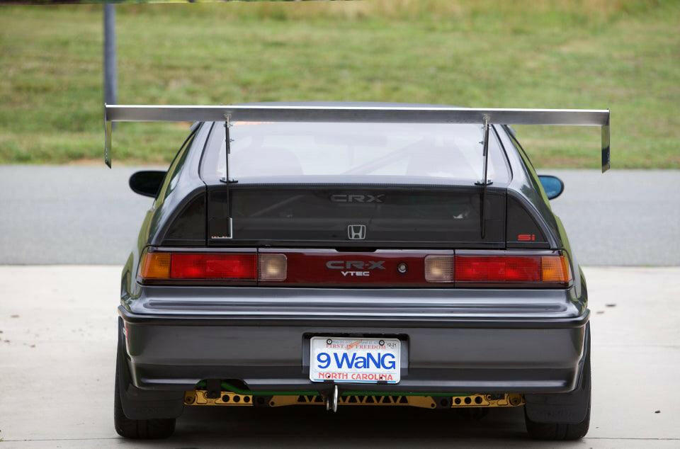A rear view of a modified dark grey Honda CRX VTEC parked on a concrete surface showcases the CRX Big Wang Kit '83-91 by 9livesracing, featuring aluminum wings for added downforce, a custom diffuser with gold accents, tinted windows, and a North Carolina license plate that reads "9 Wang." A green lawn is visible in the background.