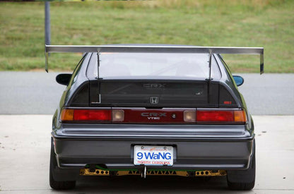 A rear view of a modified dark grey Honda CRX VTEC parked on a concrete surface showcases the CRX Big Wang Kit '83-91 by 9livesracing, featuring aluminum wings for added downforce, a custom diffuser with gold accents, tinted windows, and a North Carolina license plate that reads "9 Wang." A green lawn is visible in the background.