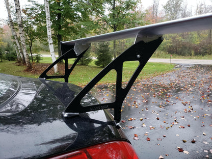 A close-up of a large rear spoiler mounted on the trunk of a black car. The 350z Big Wang Kit '02-08 Z33 by 9livesracing, crafted with extruded aluminum wings and black brackets, is set against a background of a wet driveway, fallen autumn leaves, and trees.