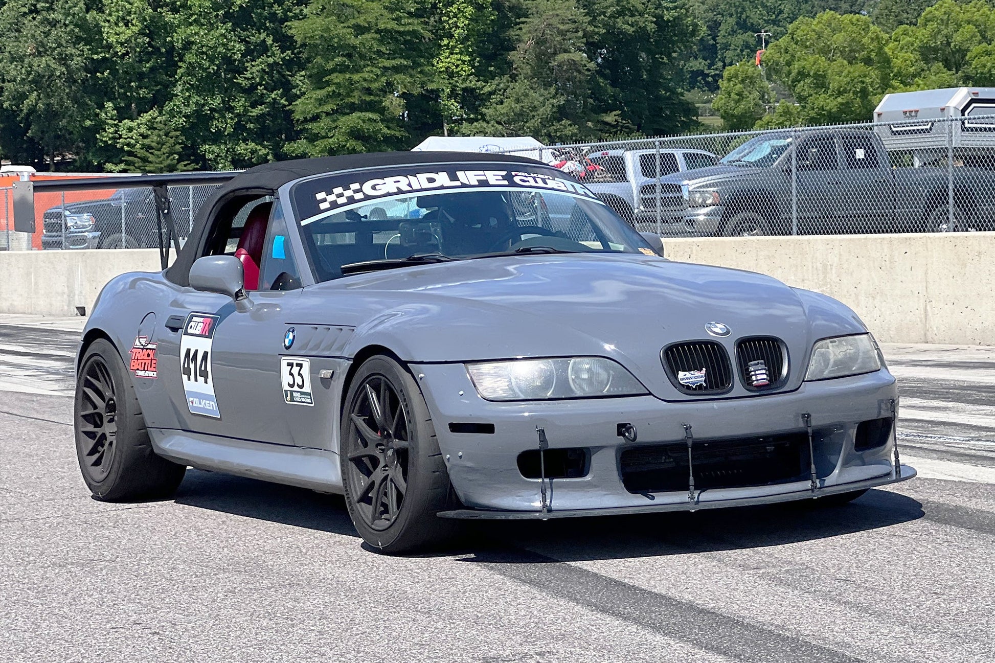 A gray BMW z3 roadster with the number 14 on the driver's door and 'Gridlife' branding on the windshield, sporting 9livesracing wings for improved lift-to-drag ratio and Big Wang GT3 performance modifications, speeds down a track with trees in the background and a barrier separating it from parked vehicles.